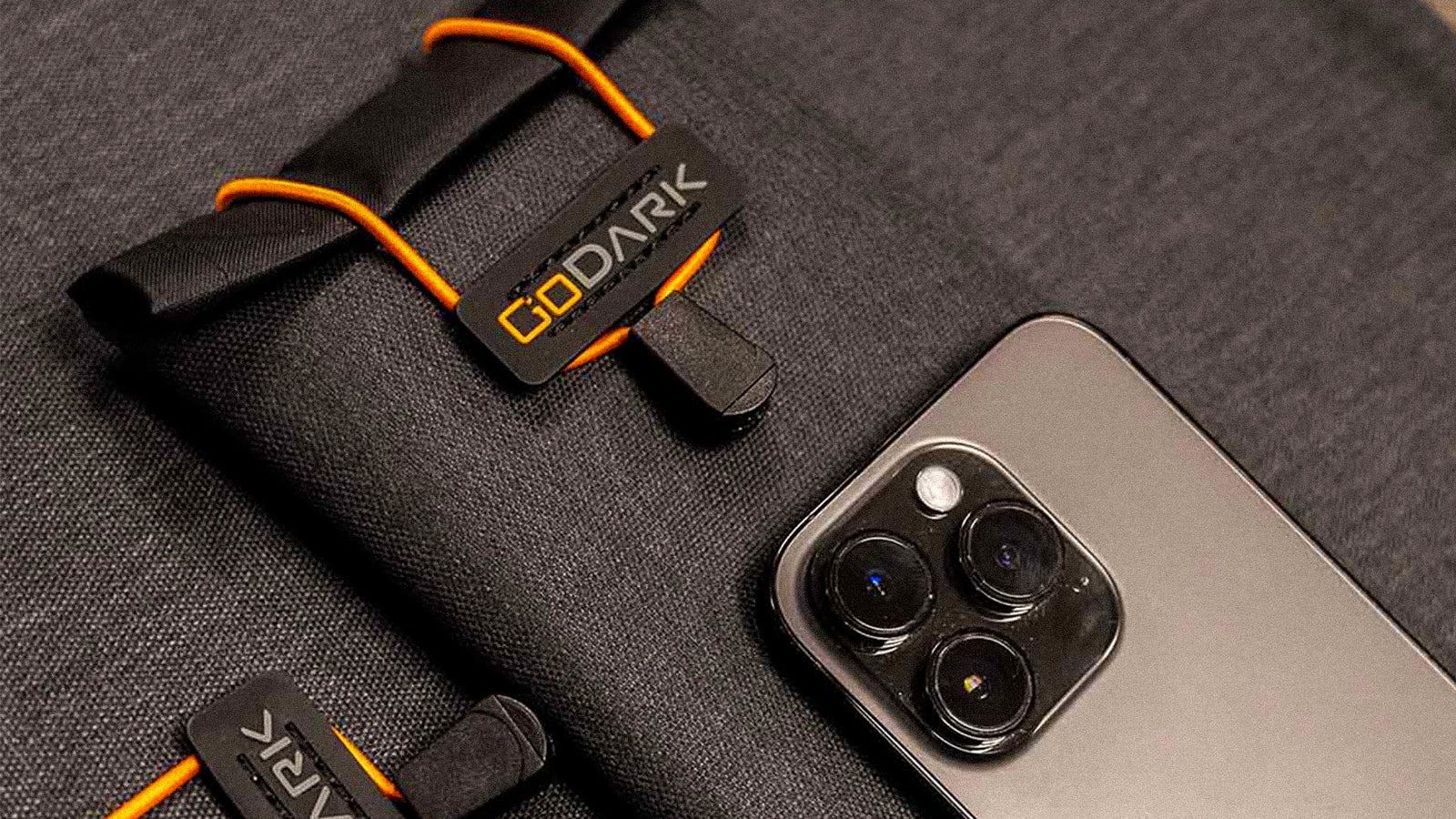 A close-up of a GoDark Faraday Bag made of black signal-blocking fabric with orange elastic cords, shown next to a smartphone with triple camera lenses, demonstrating the bag’s use for shielding devices from wireless signals.