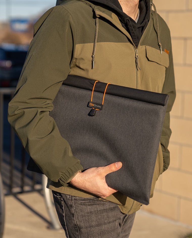 Person holding a large GoDark Laptop Sleeve with an orange strap, wearing a green jacket.